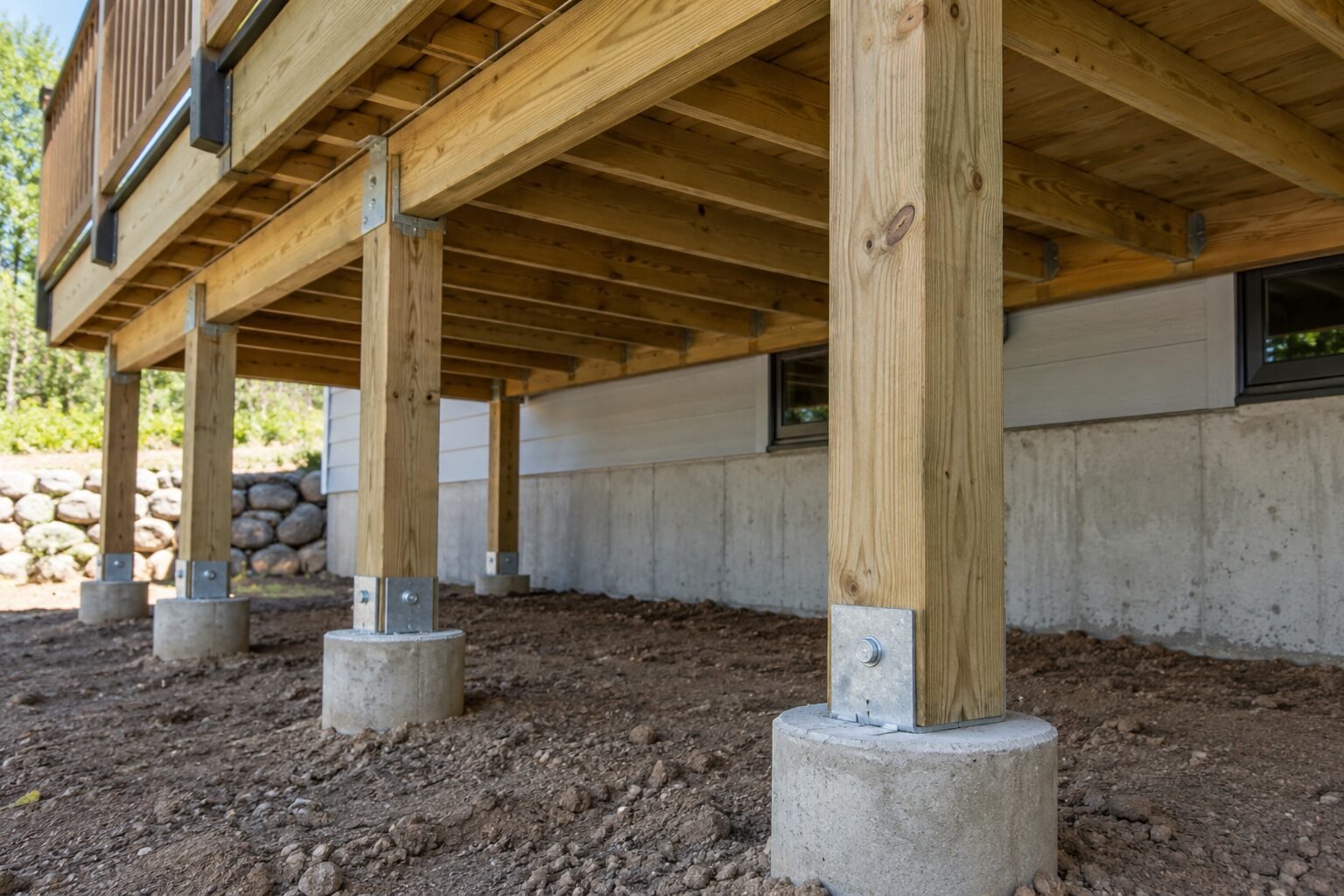 Underside of a wood deck with pressure-treated posts anchored by galvanized bases to round concrete pier footings